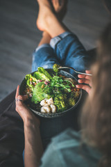 Healthy dinner or lunch. Curly woman in t-shirt and jeans sitting at home and eating vegan superbowl or Buddha bowl with hummus, vegetable, fresh salad, beans, couscous and avocado, top view - obrazy, fototapety, plakaty