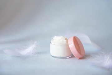 Natural face cream in a cosmetic jar on a blue background with white feathers