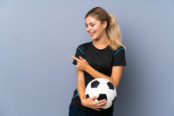Blonde football player teenager girl over grey wall pointing to the side to present a product
