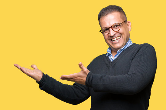 Middle age bussines arab man wearing glasses over isolated background amazed and smiling to the camera while presenting with hand and pointing with finger.