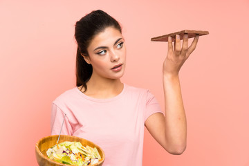 Teenager girl  over isolated pink wall with salad and chocolat and having doubts