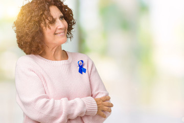 Middle ager senior woman wearing changeable blue color ribbon awareness over isolated background smiling looking to the side with arms crossed convinced and confident