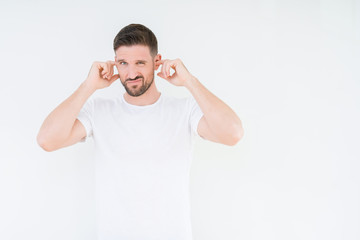 Young handsome man wearing casual white t-shirt over isolated background covering ears with fingers with annoyed expression for the noise of loud music. Deaf concept.
