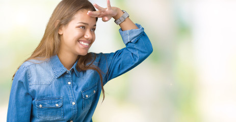 Obraz premium Young beautiful brunette woman wearing blue denim shirt over isolated background very happy and smiling looking far away with hand over head. Searching concept.