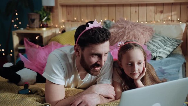 Front side view of cheerful father and girl kid dressed like fairies watching fun mobies together on laptop in the bedroom. Home family party.
