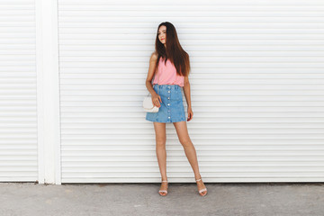 Young brunette woman wearing blue denim mini skirt, pink tank top, block heeled sandals, white cross body bag posing near white roller door. Trendy casual summer outfit. Everyday look. Street fashion.
