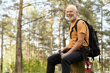 Naklejka na ściany i meble People, adventure, travel and active healthy lifestyle concept. Cheerful energetic elderly man hiking with backpack in forest, having rest on stump, drinking water with pine trees in background