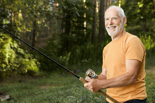 Summertime image of skilled fisherman on retirement having rest in wild nature using fishing rod, waiting for fish to be caught, looking at camera with smile. Mature people, leisure and active hobby