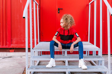 fashionable teenager girl posing on a red wall background, sitting on the stairs