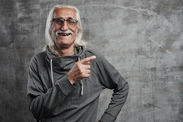 Aged gray haired smiling Caucasian man points to the side with index finger, advertises something. Grandfather in glasses with mustache recommends buying your product