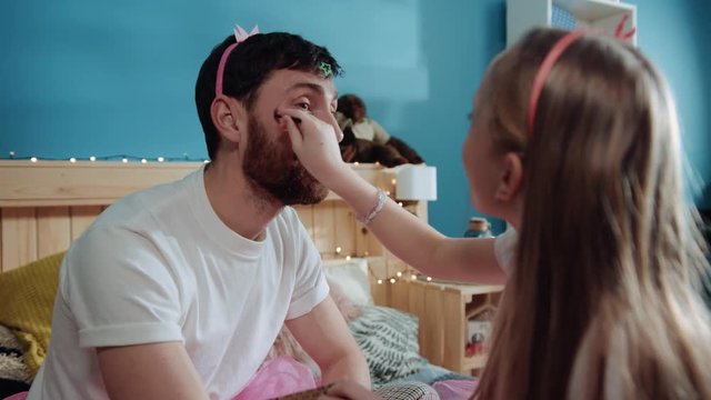 Handsome young man plays with her daughter while sitting on bed. Little preteen fairy girl applying make-up of her daddy's face and laughing together.