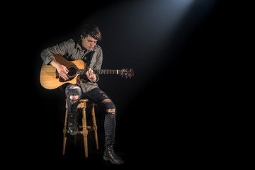 Fototapeta premium musician playing acoustic guitar, sitting on high chair, black background with beautiful soft light