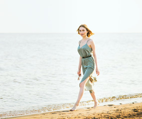 Young beautiful slim woman with short light hair wearing green summer dress walking at sea coast at sunset.