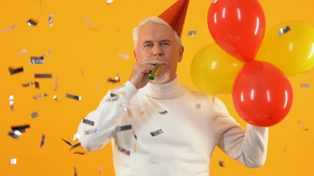 Mature male party hat blowing noisemaker and holding balloons, birthday greeting