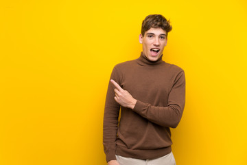 Handsome young man over isolated yellow background surprised and pointing side