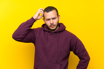 Colombian man with sweatshirt over yellow wall having doubts while scratching head