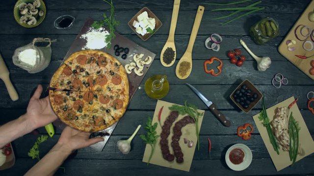Pizza on ecological black background.  The cook puts finished pizza on table. Meat pizza with different filling: salami, mushrooms, sausage, cheese, olives. There are also many other products on table