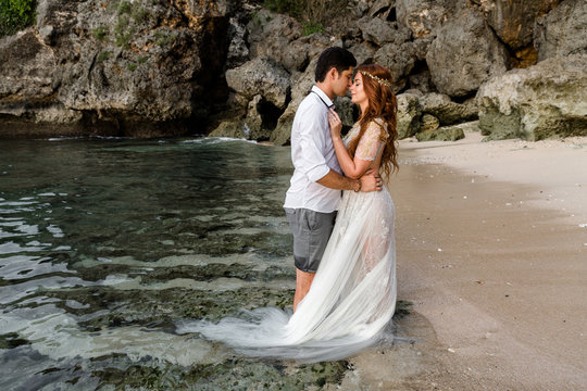 Bride and groom embrace standing in the sea water on the beach against the backdrop of the cliff. Young couple bowed to each other before a kiss. Newlyweds at wedding day on ocean beach