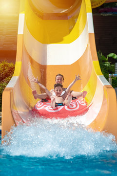 European boy and mom gliding down slide in waterpark. They enjoy the fun and holding hands wide open.