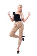Playful blonde fashion model woman posing on one leg showing two finger victory gesture.  Full body isolated on white background.