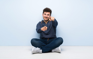 Young man sitting on the floor points finger at you while smiling
