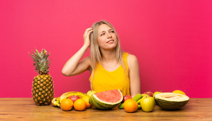Young blonde woman with lots of fruits having doubts and with confuse face expression