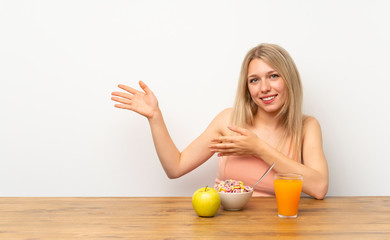 Young blonde woman having breakfast extending hands to the side for inviting to come