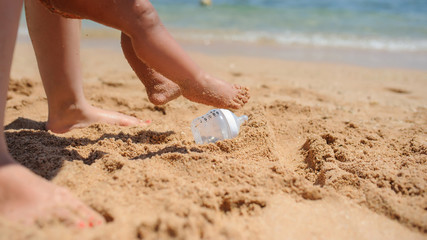 close up photo of a little girl legs in swimsuit walking on the beach with her mother and a bottle for milk on the sand