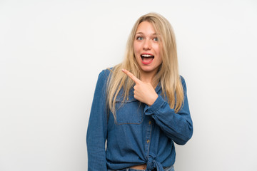 Young blonde woman over isolated white wall surprised and pointing side