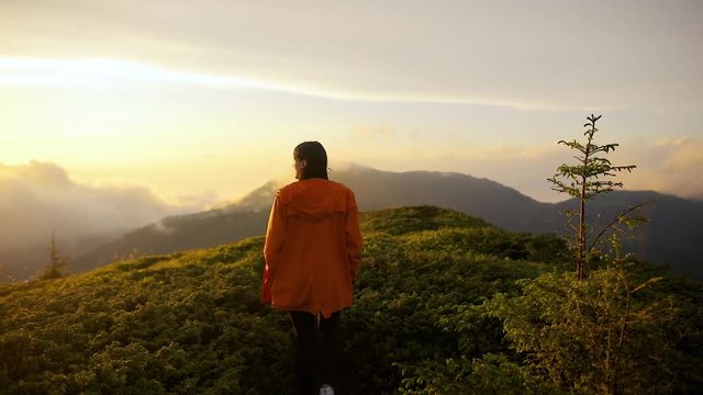 Camera follows hipster millennial young woman in orange jacket running up on top of mountain summit at sunset, happy and drunk on life, youth and happiness