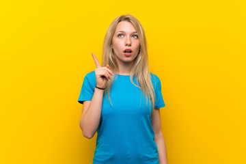 Young blonde woman over isolated yellow background thinking an idea pointing the finger up
