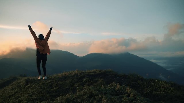 Camera follows hipster millennial young woman in orange jacket running up on top of mountain summit at sunset, jumps on top of rocks, raises arms into air, happy and drunk on life, youth and happiness