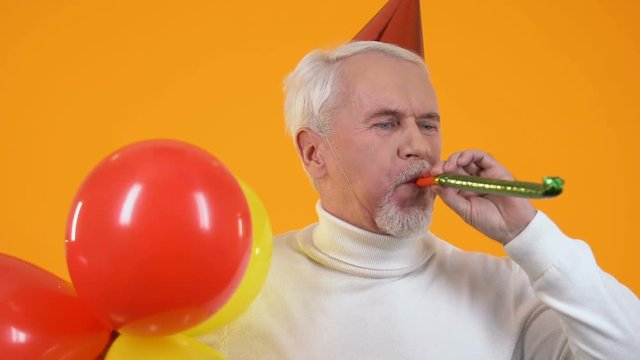 Joyful senior man party hat blowing noisemaker holding balloons, celebration