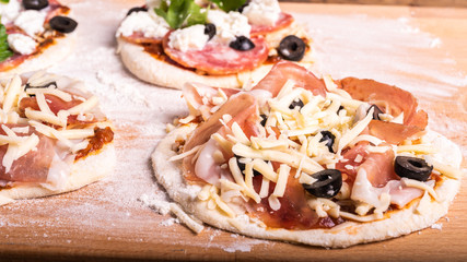 Cooking traditional Italian mini pizza, pizzetta - raw mini pizza with tomato sauce, black olives, jamon and mozzarella cheese on a cutting board, close-up