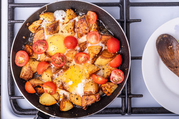 Men's rustic breakfast - fried potatoes with eggs and cherry tomatoes in a pan, standing on the stove