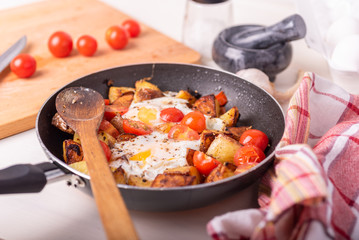 Men's rustic breakfast - fried potatoes with eggs and cherry tomatoes in a pan, standing on a wooden kitchen table.
