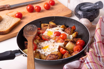 Men's rustic breakfast - fried potatoes with eggs and cherry tomatoes in a pan, standing on a wooden kitchen table.