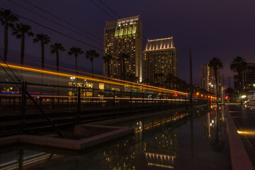 Fototapeta na wymiar Streaks of Light from a Passing Trolley in the City of San Diego, California at Night - with City Lights Reflected in the Water, and Rows of Buildings and Palm Trees in the Background