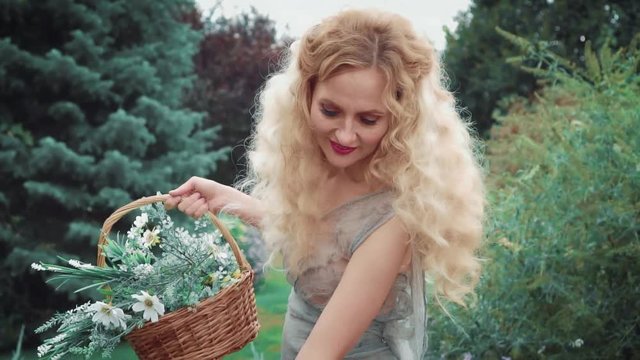 pretty attractive woman with lush blond curly hair gathers summer purple flowers, kind witch smiles and strokes wonderful magical healing herbs, happy sorceress collects the ingredients for potion