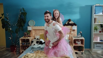 Two pretty princesses father and daughter playing together at home party. Happy daddy holding young girl on his back doing tricks with magic wand on bed.