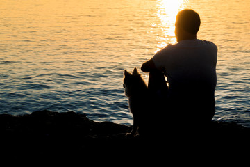 Silhouette of man and dog sitting together at the seaside and looking at the sunrise