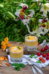 Two galasses of peach panna cotta with white and orange layers. Served outdoor on wooden background with mint and cherry.