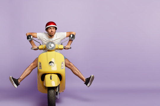 Funny young man with fasten motorbike helmet, poses on fast bike, wears white t shirt and sneakers, poses against purple background with empty space. People, transportation and riding concept
