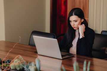 Young attractive emotional girl in business-style clothes sitting at a desk on a laptop and phone in the office or auditorium
