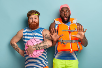 Friendly multiethnic guys with swimming equipment, ready for summer vacation. Scared dark skinned man in swimcap and lifejacket shows refusal gesture, red haired friend in sailor vest with beach ball