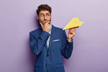 Contemplative handsome businessman holds chin, holds yellow paper plane, thinks over successful...