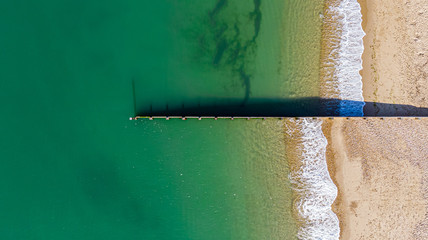 An aerial view of a groyne (breakwater) dividing a majestic cystal clear green water sea with crashing waves on a sunny sandy beach