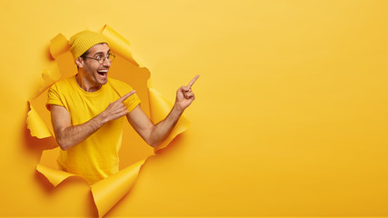 Pleased cheerful man attracts your attention to special offer, points at upper right corner, poses in torn paper hole of yellow background. People, promotion concept. Satisfied guy promots object - obrazy, fototapety, plakaty