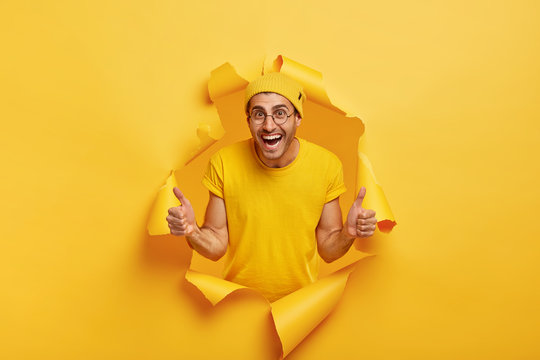 Young cheerful young male model gives thumb up gesture, stands through paper hole, smiles broadly, wears yellow hat and t shirt, demonstrates approval and agreement, agrees with awesome idea