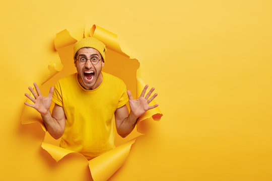 Emotional unshaven guy stands in ripped paper hole of yellow background, yells loudly, gestures and shows palms, tries to prevent you from danger, shouts be carefully, wears bright hat and t shirt